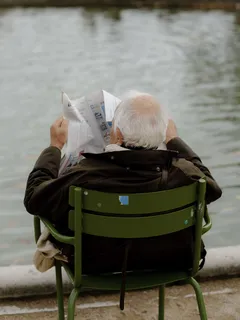 Person reading newspaper on bench by water under sunny sky